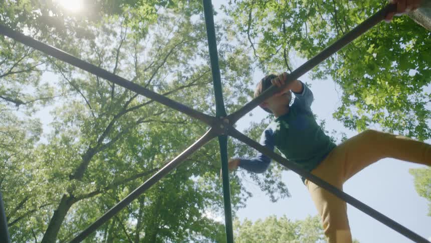 Boy with blue sweatshirt and yellow pants climbs monkey bars at the playground