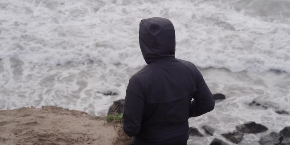 Hooded person walking down an incline towards a rocky beach