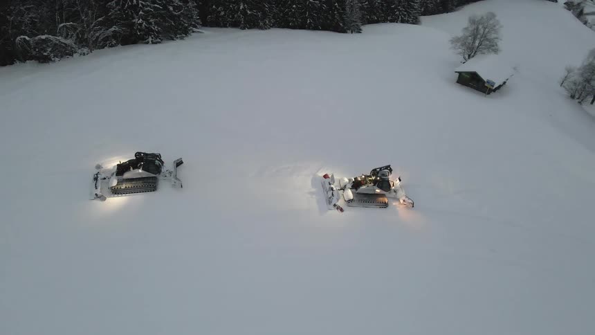 Two large snow plows move steadily through thick snow on their way towards a remote group of houses