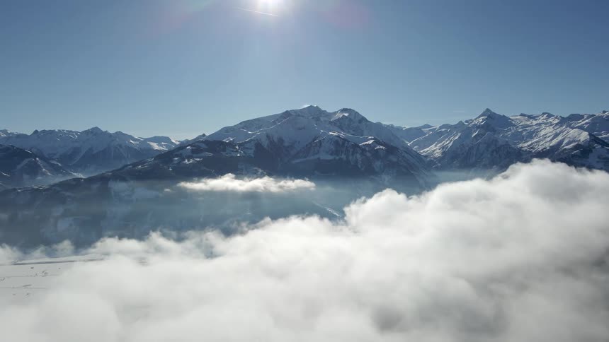 Snow covered mountains high in the clouds with a bright sun flare shining in a blue sky