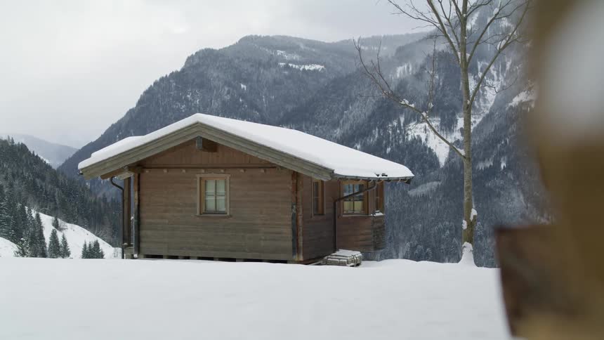 A cabin covered in snow next to a tree with a mountain range the sky and clouds in the background