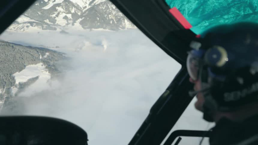 A man with goggles and a helmet in the cockpit of an aircraft with a mountain range the sky and clouds in the background