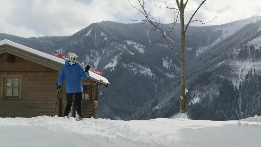 A man walks uphill through thick snow towards a cabin while holding his skis atop his shoulder