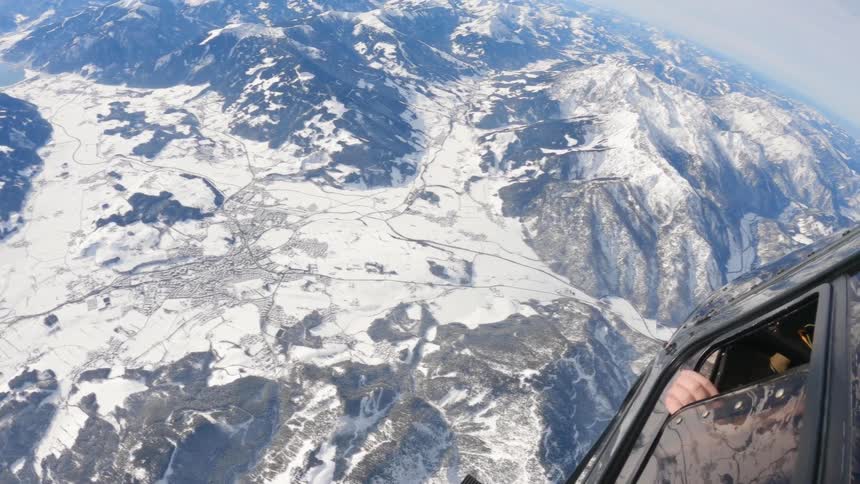 A parachuter clinging to the side of a helicopter flying over snowy mountains