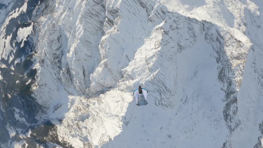 A man jumps out of a helicopter and glides down in an air gliding suit above a snowy mountain top at daytime
