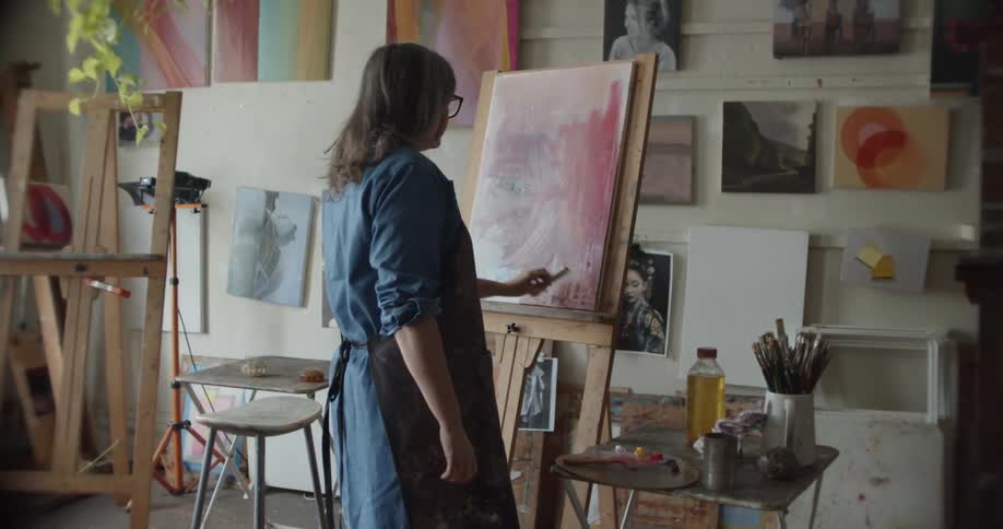 Woman wearing an apron painting with a small brush over canvas on an easel inside a studio takes a picture of a painting