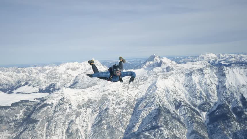 A man with a helmet skydiving with a mountain range the sky and clouds in the background