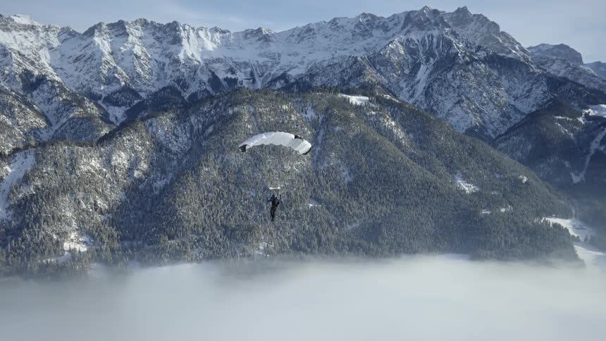 A man paraglides towards a snowy mountain top with trees on its slope during the day