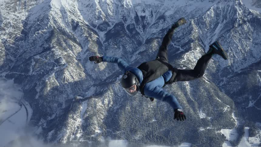 Skydiver wearing a parachute pack and black helmet is floating over snow covered mountains