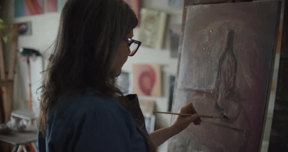 Woman wearing an apron painting with a small brush over canvas on an easel inside a studio