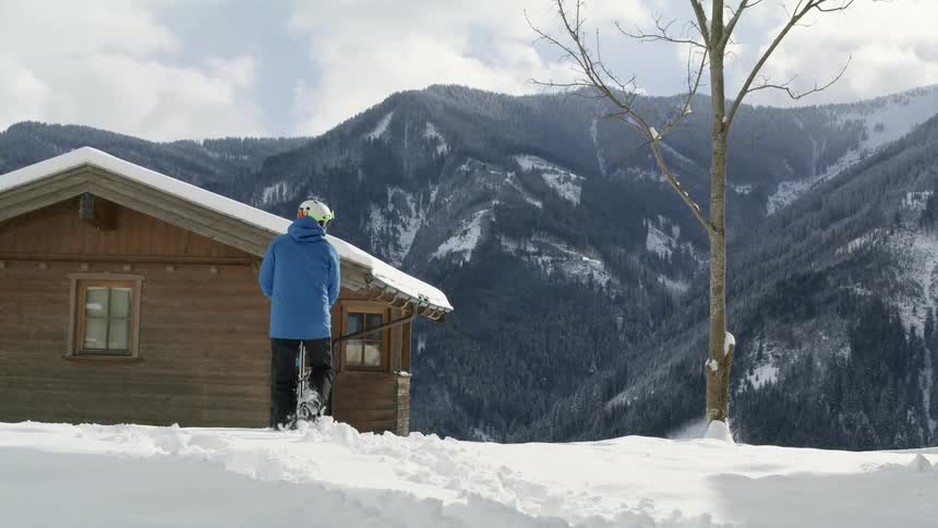 A person holding skis walks by a cabin in the mountains