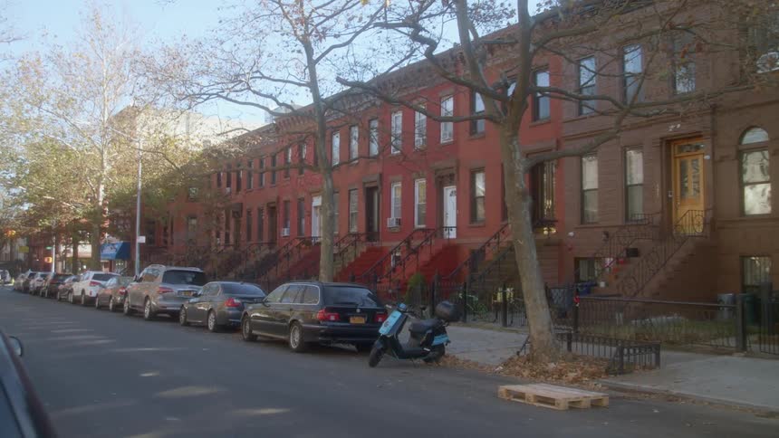 A city street with brownstone houses with vehicles trees and with the sky in the background