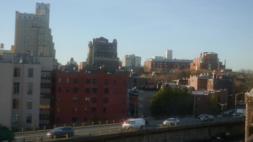 Cars and trucks travel on a city highway past buildings on a sunny day