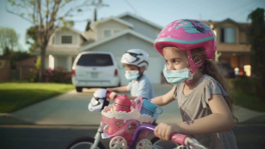 A boy and girl ride their bicycles past neighborhood homes and driveways while wearing helmets and masks