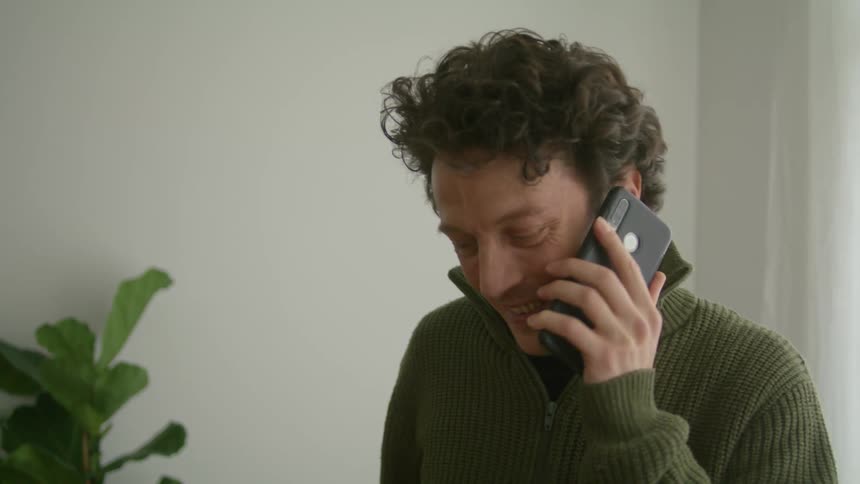 A man standing indoors smiles as he speaks on his smartphone before removing the phone from his ear