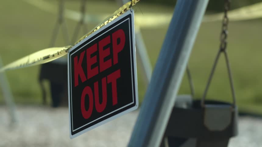 A keep out sign hangs from yellow caution tape blocking access to swings at a playground
