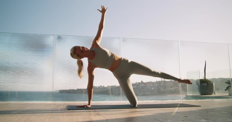 A young woman does yoga exercises on a mat on a balcony overlooking the coast of a city on a sunny day