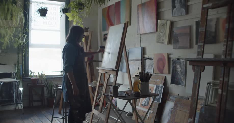 A woman adds more paint to a canvas on an easel in a room filled with artwork