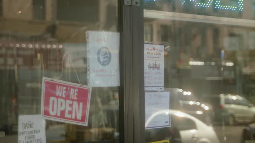Signs hang in the window of a business with cars parked outside and people passing by