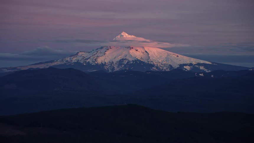 A mountain covered in snow and ice with the sky and clouds in the background as the camera slowly zooms out showing the vastness of the forest in Oregon