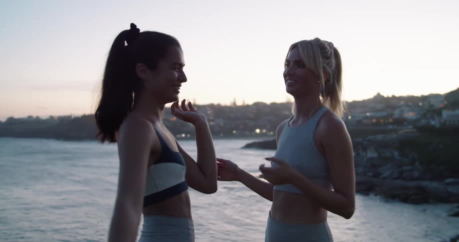 Two woman smile and laughing as they stand outside near a scenic shoreline