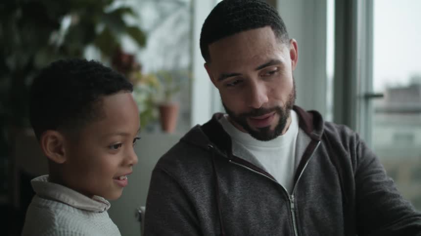 A father talking to his young son while working on a laptop at home