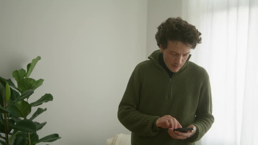 A middle aged man taps the screen of his smartphone as he stands indoors near a window during the day