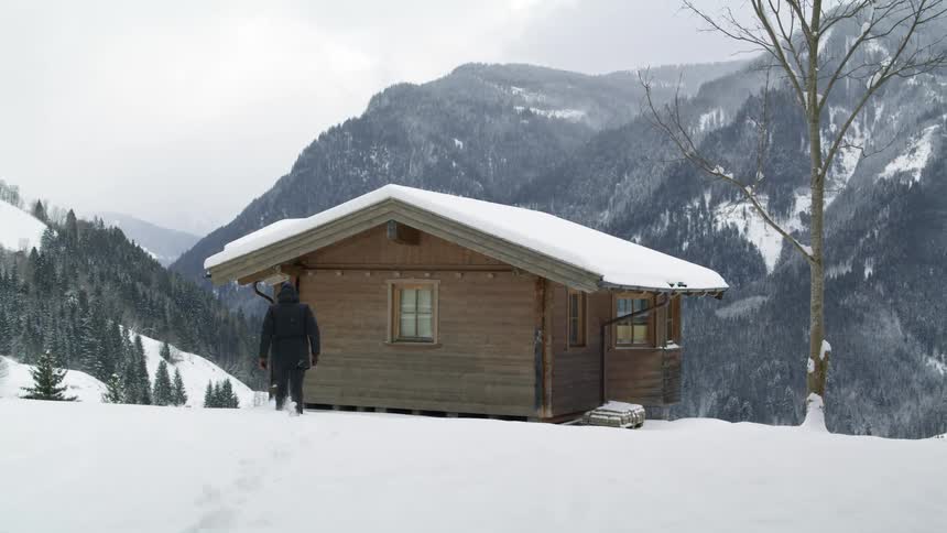 A man walks towards a log cabin on top of a snowy mountain next to a mountain range during the day