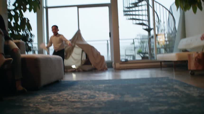 A little boy running around in a living room playing with a paper airplane during the day