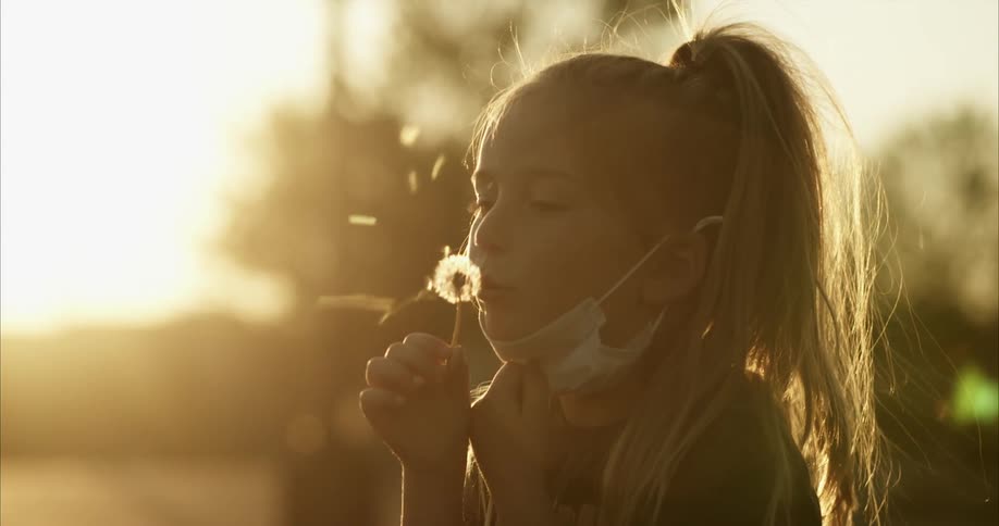Young girl pulls down her face mask to on a dandelion flower at sunset