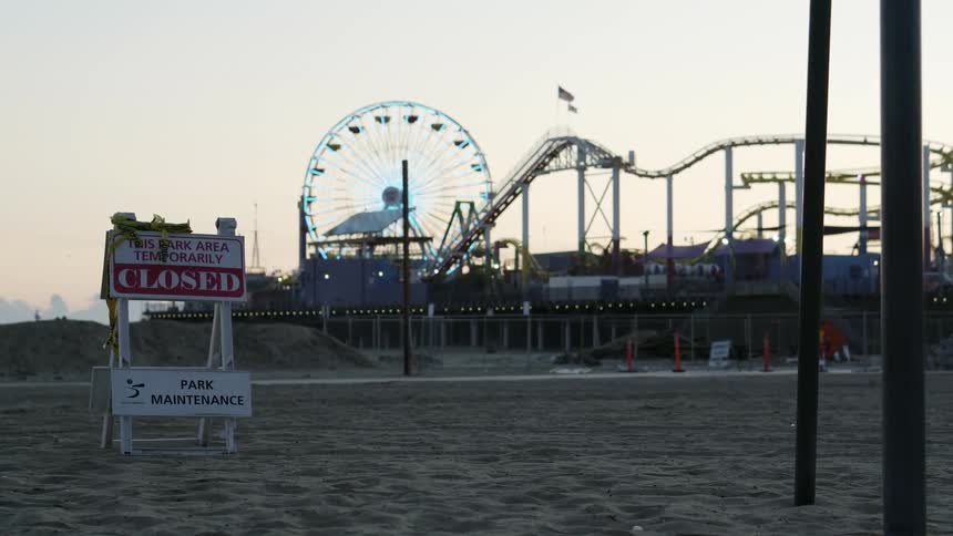 A closed sign stands in the sand in front of an amusement park with ferris wheel and roller coaster