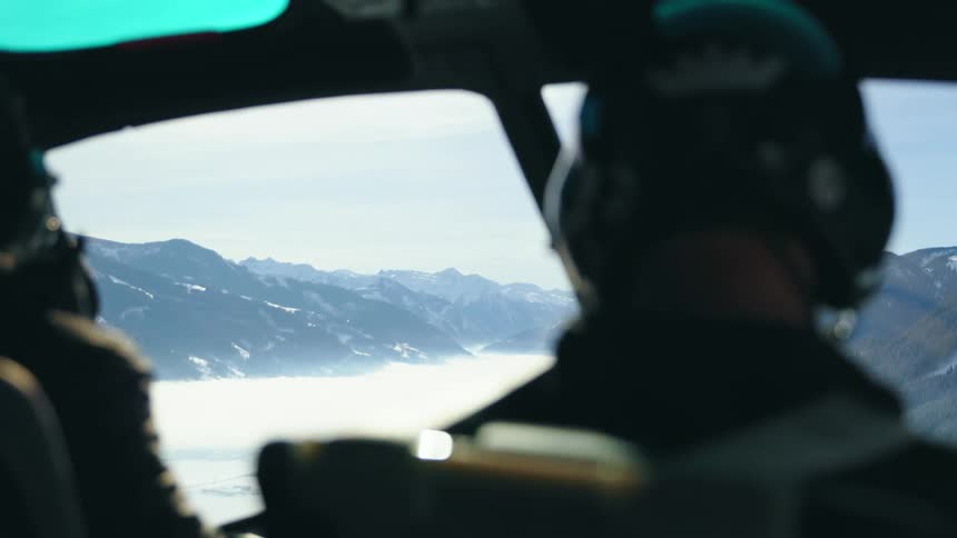 Two men piloting a helicopter flying over a snowy mountain range during the day