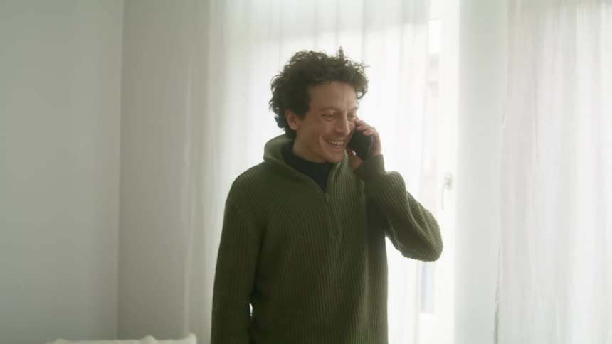 Young man wearing a green hoodiee answering a call on his smartphone at home