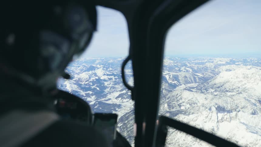 A person with a helmet in the cockpit of a helicopter with a mountain range the sky and clouds in the background