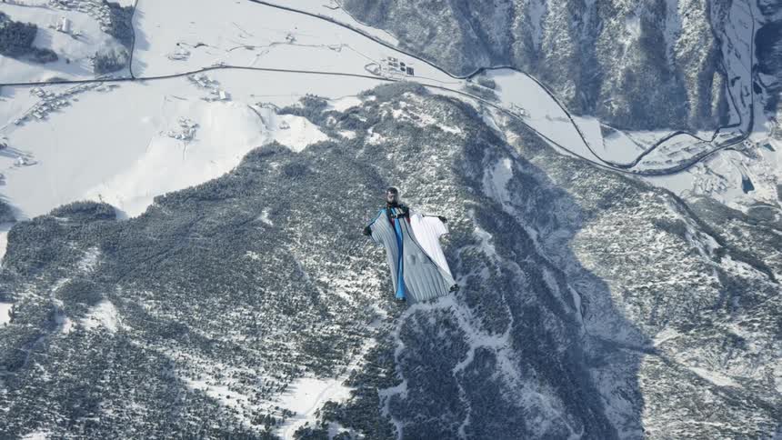 A person in a wingsuit gliding over a forested and snowy mountain range while facing downward turns to face upwards toward the camera