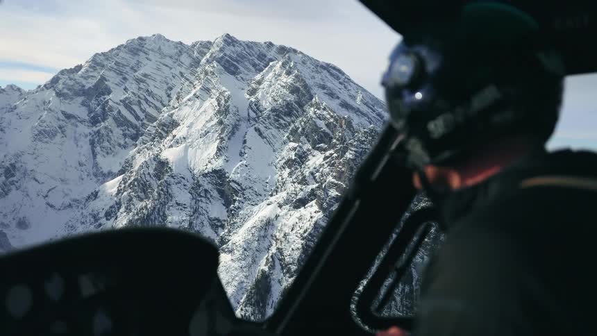 Pilot wearing a helmet piloting a helicopter over the snow covered peaks of a mountain range