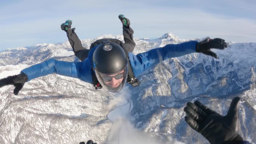 A man skydiving over the snow covered peaks of a mountain range reachs to hold the hands of the cameraman