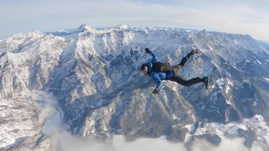 Skydiver wearing a parachute pack and black helmet is floating over a snow covered mountain canyon