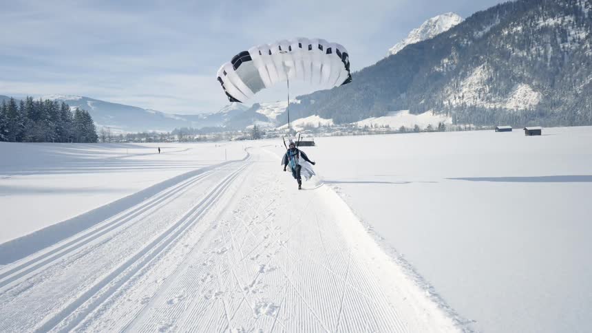 A person paragliding and landing on the snow with a mountain range the sky and clouds in the background