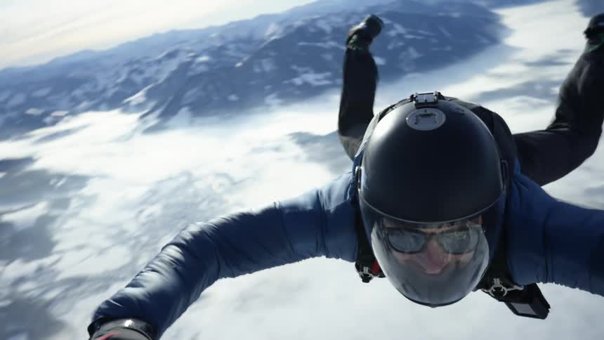 Skydiver wearing a parachute pack and black helmet with sun glasses is floating over snow covered mountains
