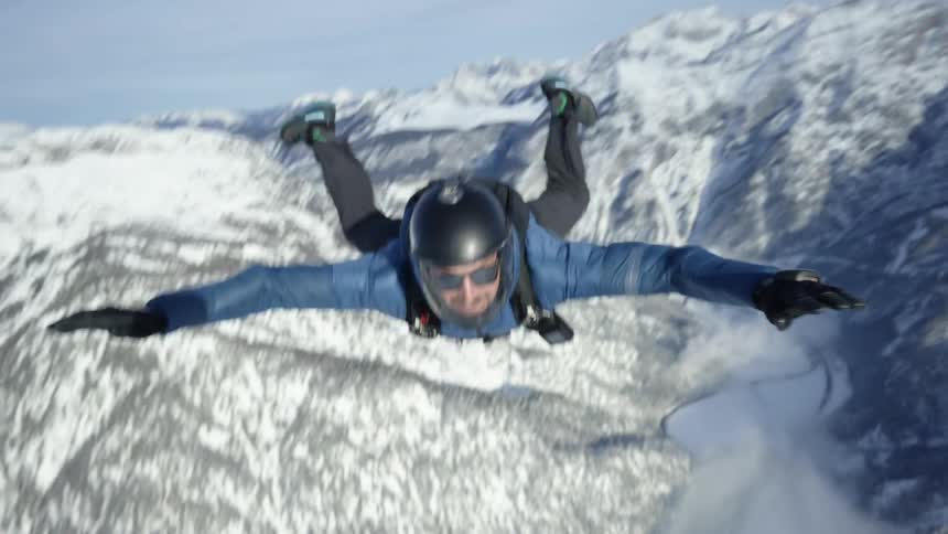 Skydiver wearing a parachute pack and black helmet with sun glasses is floating over snow covered mountains