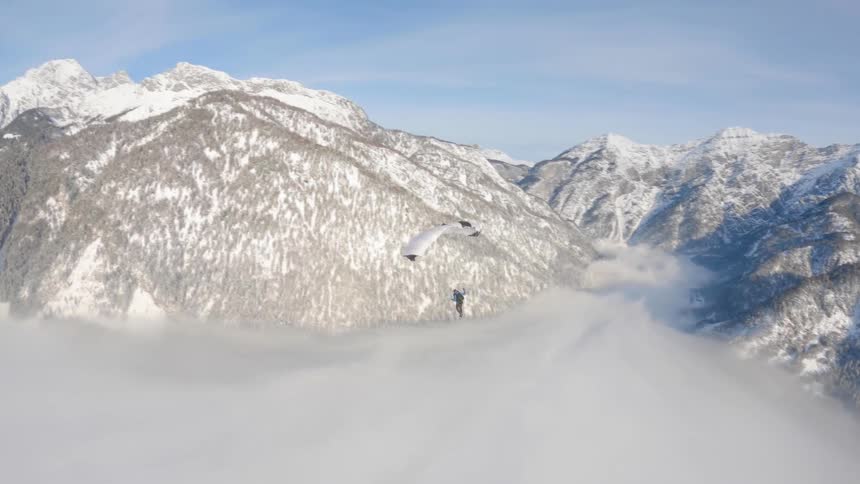 Person wearing a parachute is gliding over a foggy snow covered mountain canyon