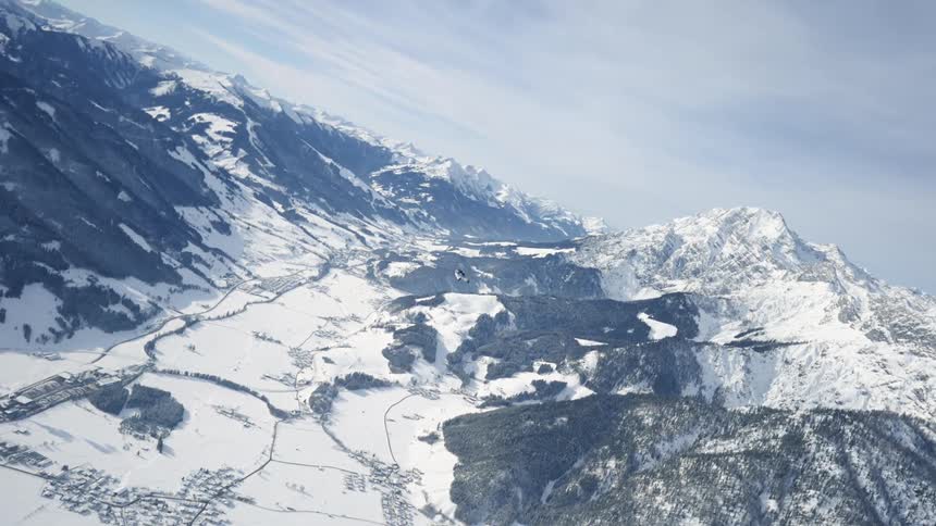 Skydiver who is wearing a wing suit is floating and gliding over a snow covered mountain canyon as they open their parachute and continue to glide through the air