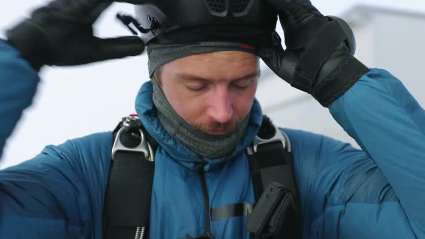 A man standing in front of an airplane hangar putting on helmet and wearing a parachute