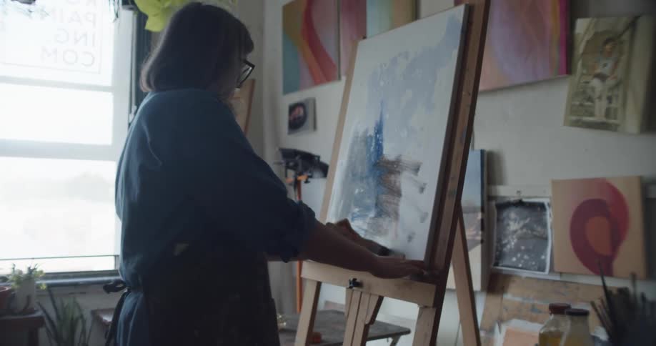 An older woman painting on a canvas in a studio with artwork and plants