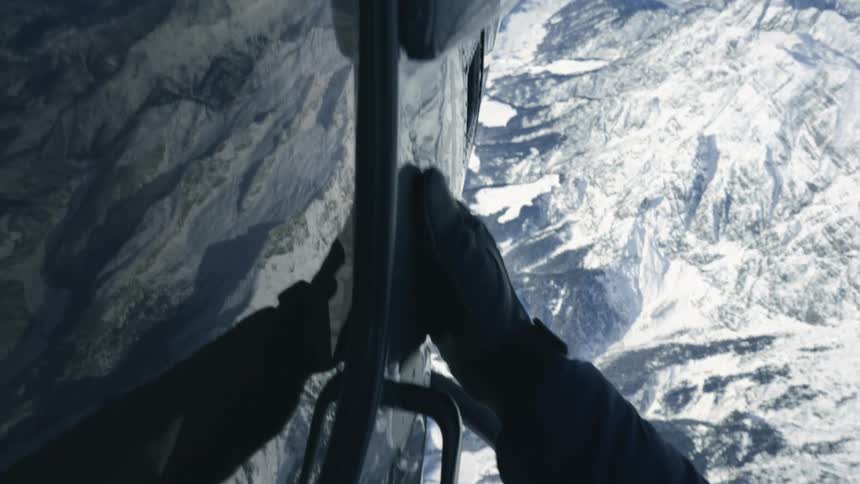 A person clinging to the side of a helicopter while flying over snowy mountains