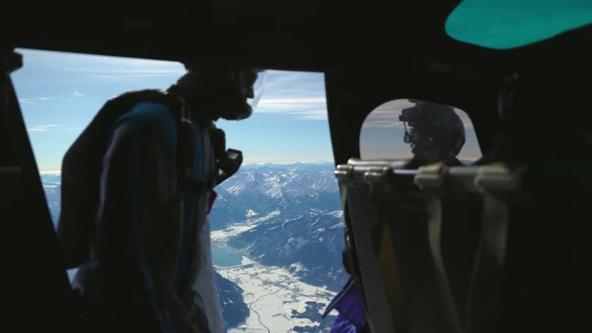 A man wearing parachuting gear stands at the entrance of a helicopter opening in the air over a snowy mountain range
