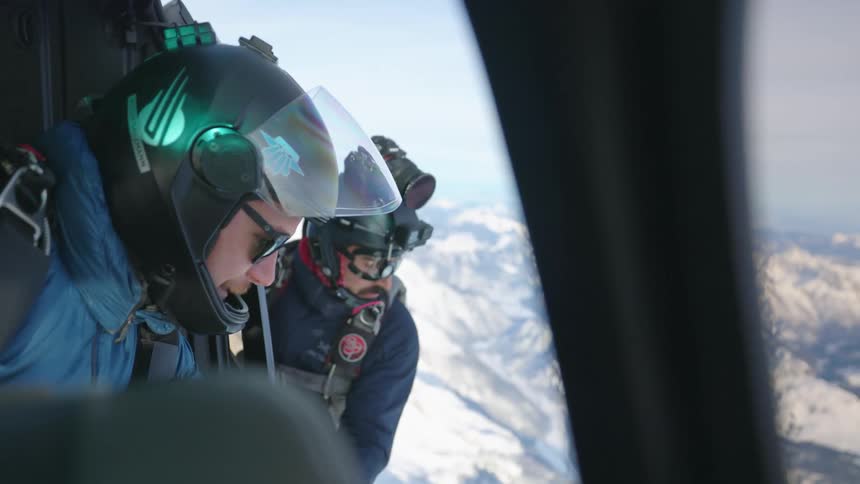 Two men with helmets on a helicopter with a mountain range the sky and clouds in the background