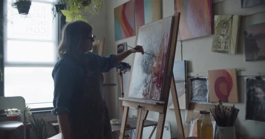 Woman wearing an apron painting with a small brush over canvas on an easel inside a studio