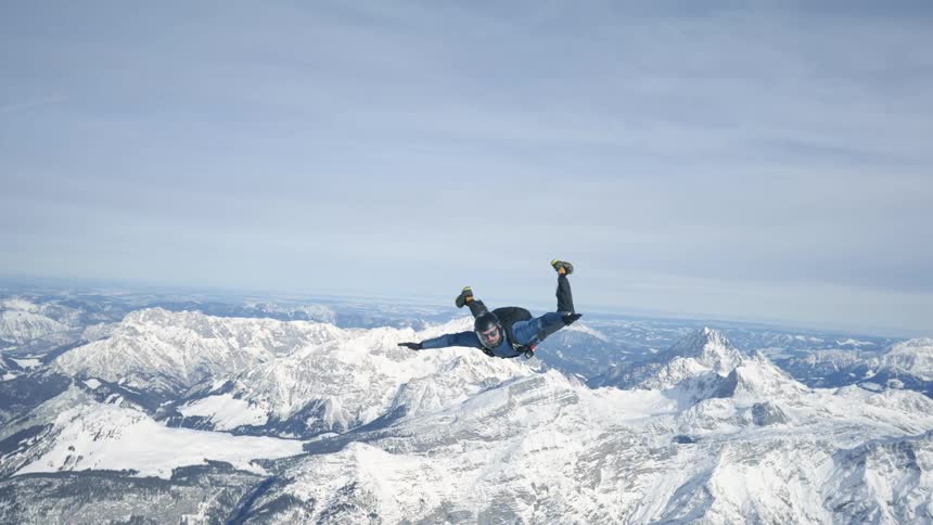Skydiver wearing a parachute pack and a black helmet is floating over snow covered mountains